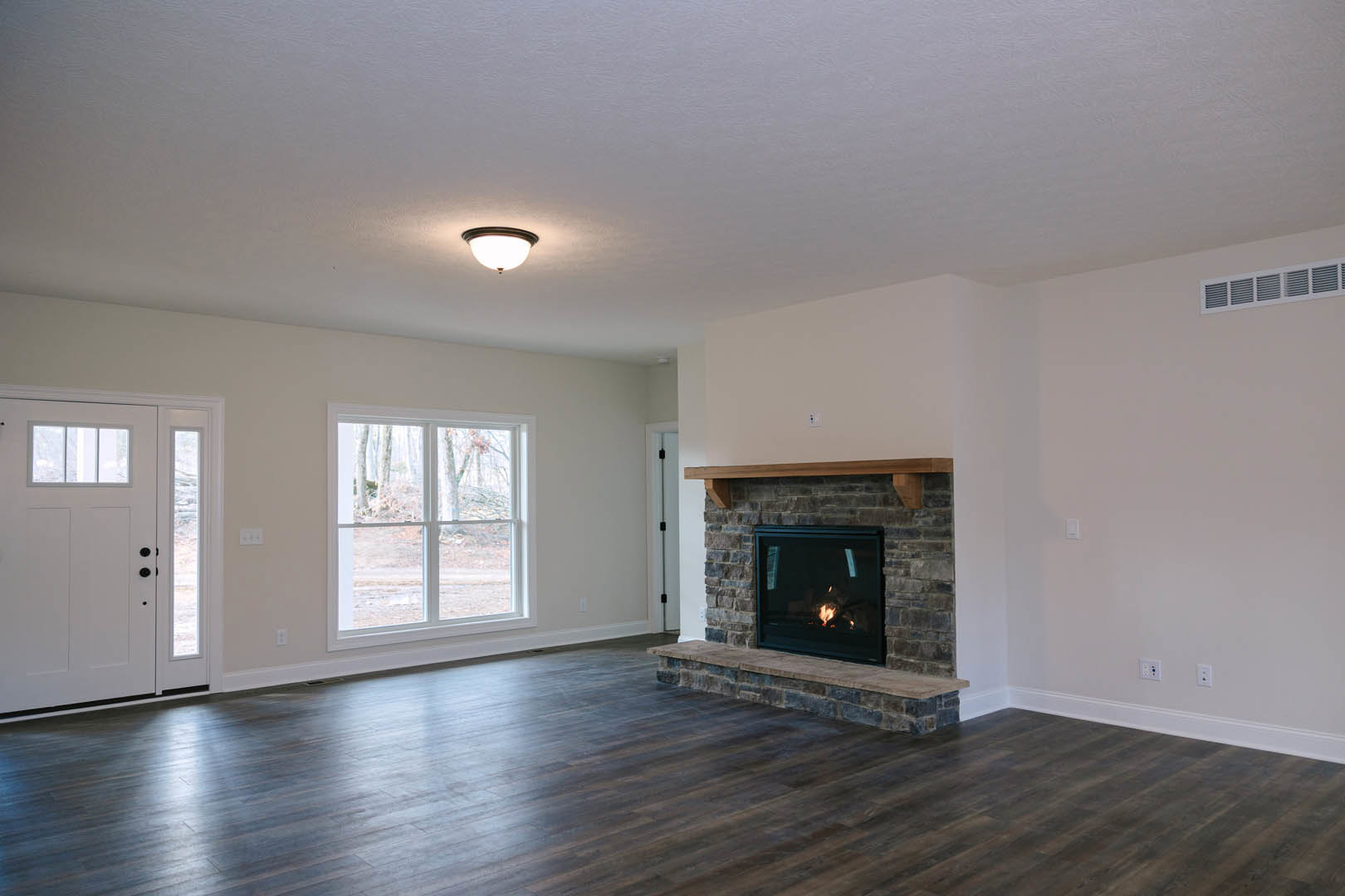 Living room with hardwood floors, stone fireplace with burning fire, large window showing trees, white door with glass panels, recessed ceiling lights