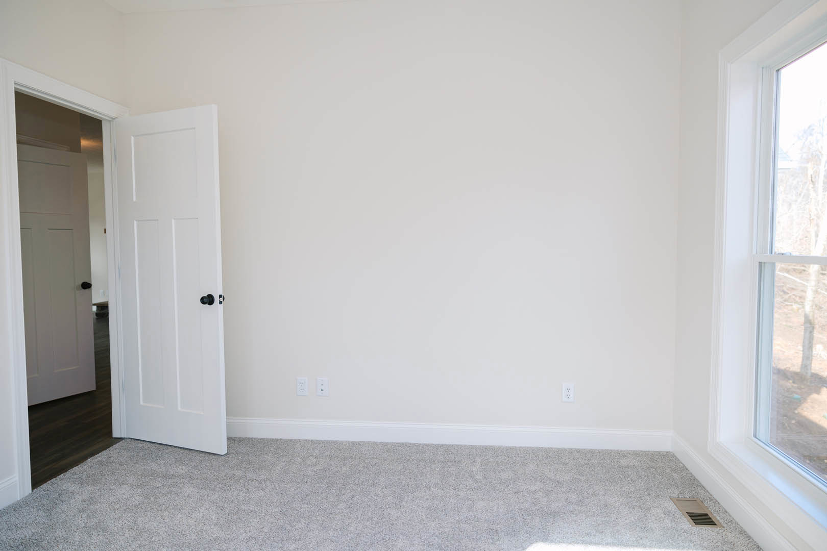 White walls and ceiling, open white door, grey carpet flooring, window with white trim, wall vent visible.