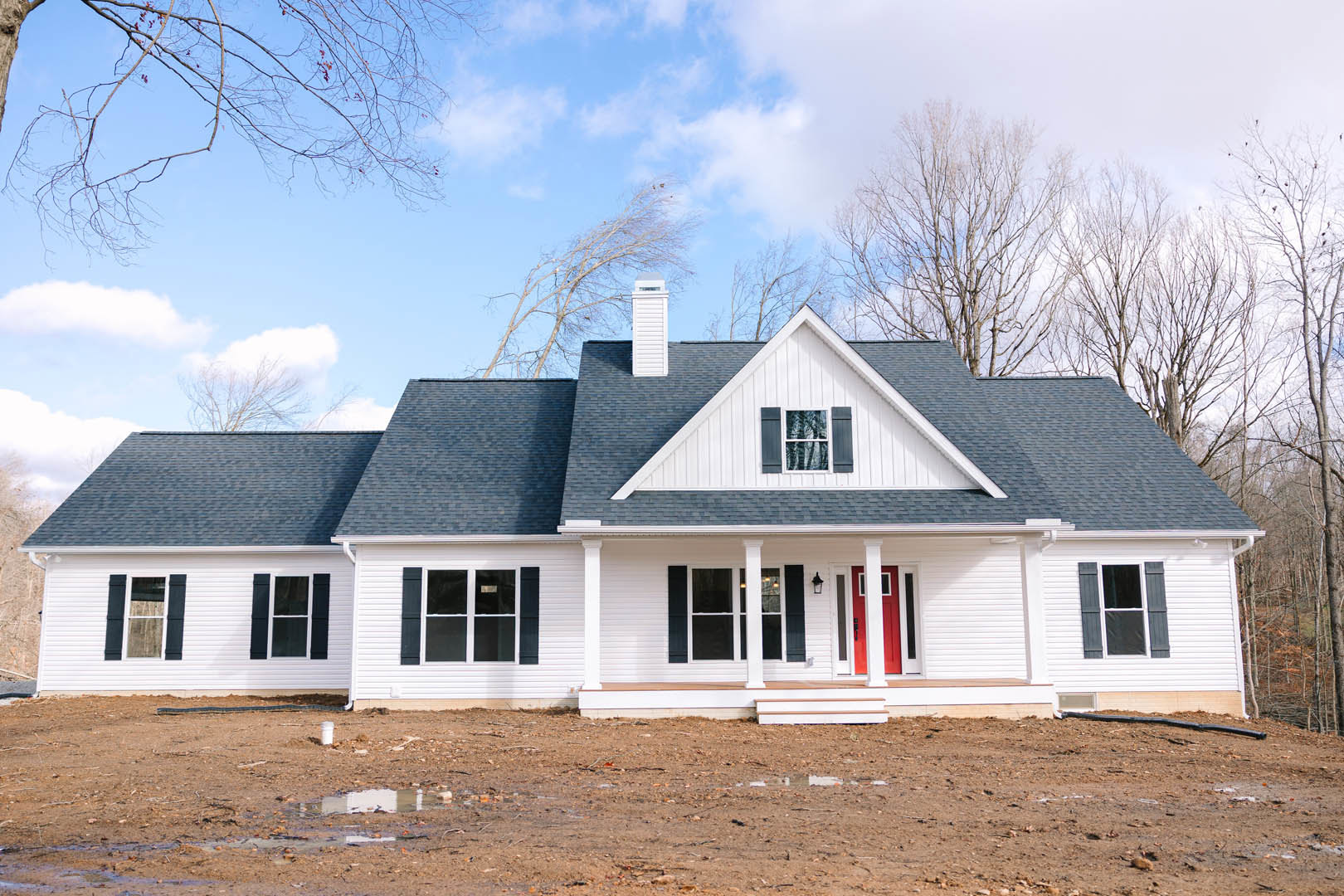 White siding house with black shutters, red front door, white-framed windows, horizontal wall panels, dirt yard, and entry stairs under a cloudy sky.