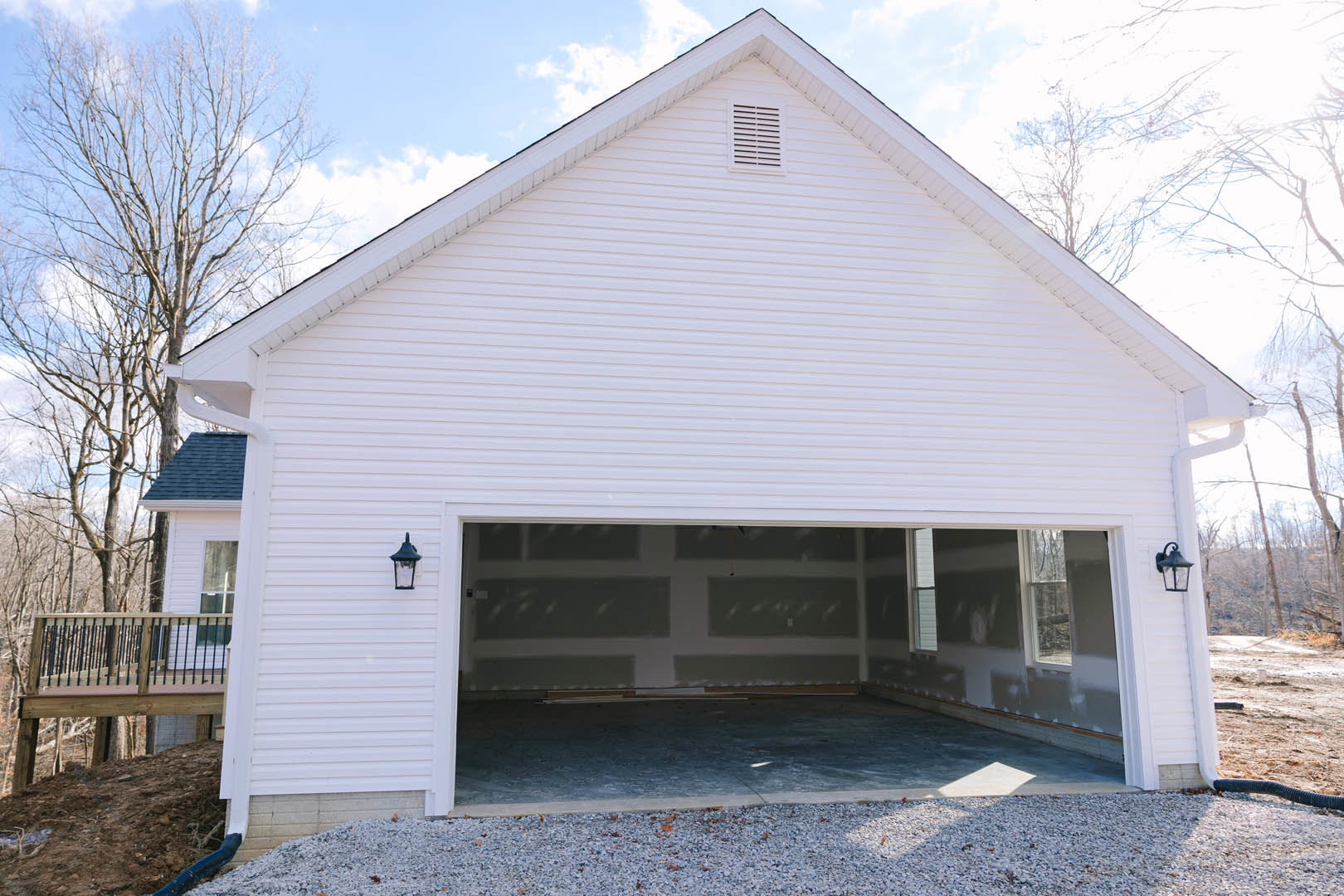 Detached garage with white metal roof, gray siding, exterior wall lamp, black railing on adjacent wooden deck, gravel driveway, and surrounding trees under cloudy sky