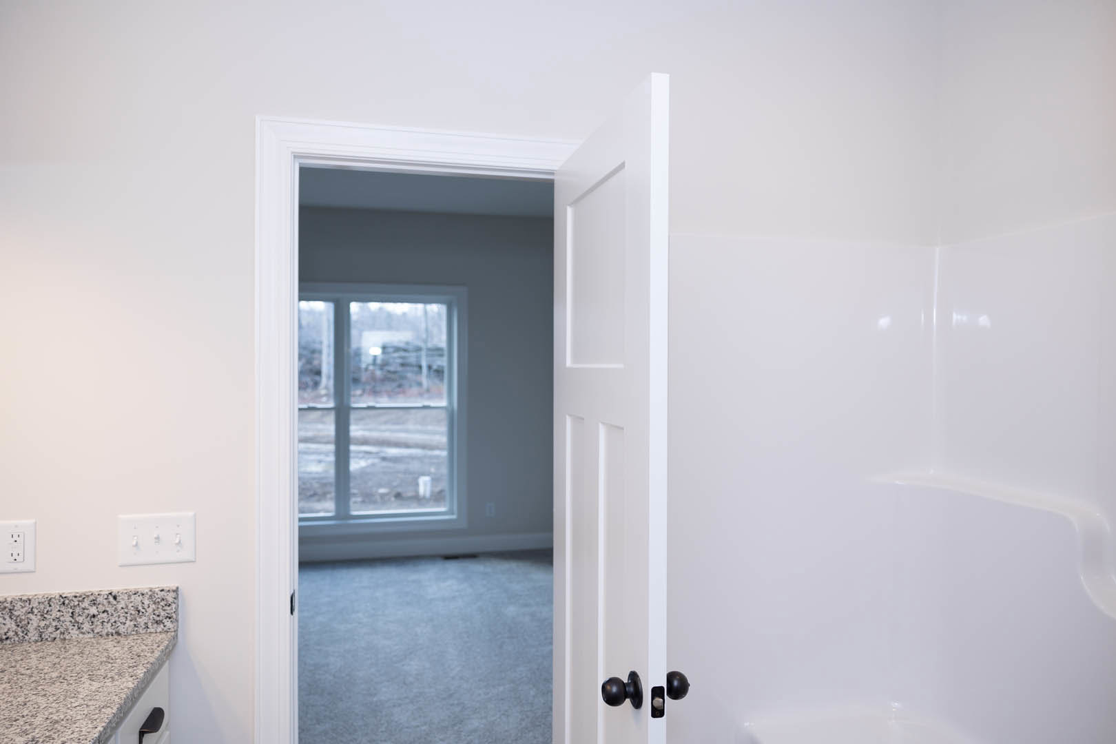 White door open to a bathroom with tile flooring, bathtub, sink, and chrome plumbing fixtures; visible countertop, carpet, window, and white switch with knobs.