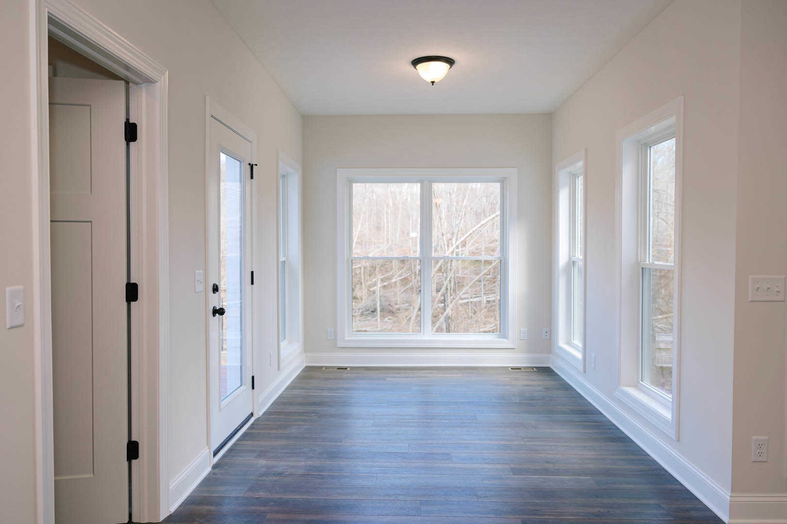 Hallway with large windows, natural light, blue-toned wood flooring, white trim, modern light fixture, white door, and wall switch; trees visible outside