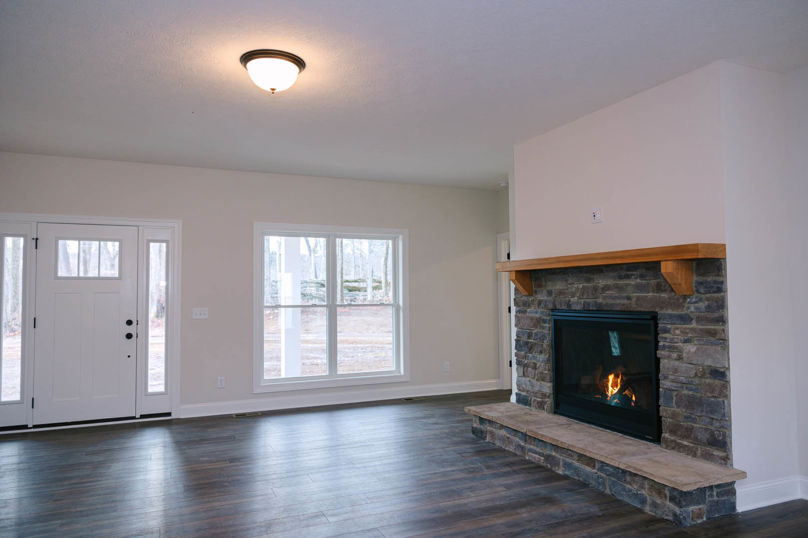 Stone fireplace with burning fire, wood flooring, ceiling light fixture, white door with glass panes, large window showing outdoor view