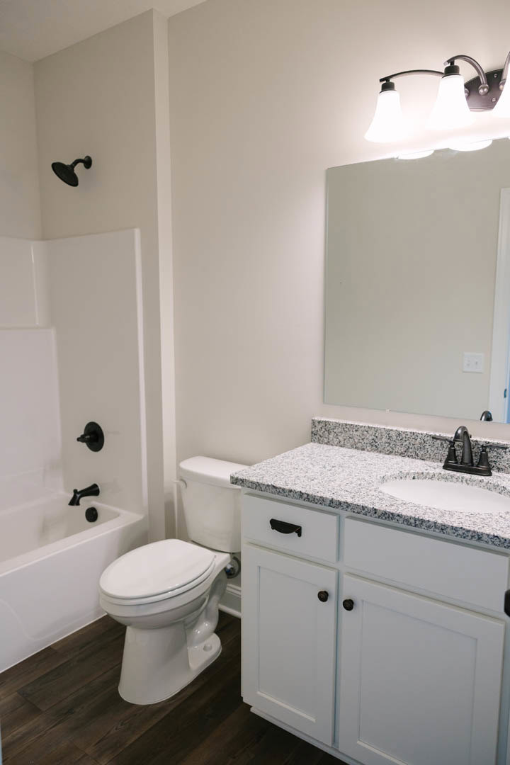 Bathroom with marbled countertop, black faucet, white sink, toilet with white lid, freestanding bathtub, black shower head, light fixture, and tiled walls