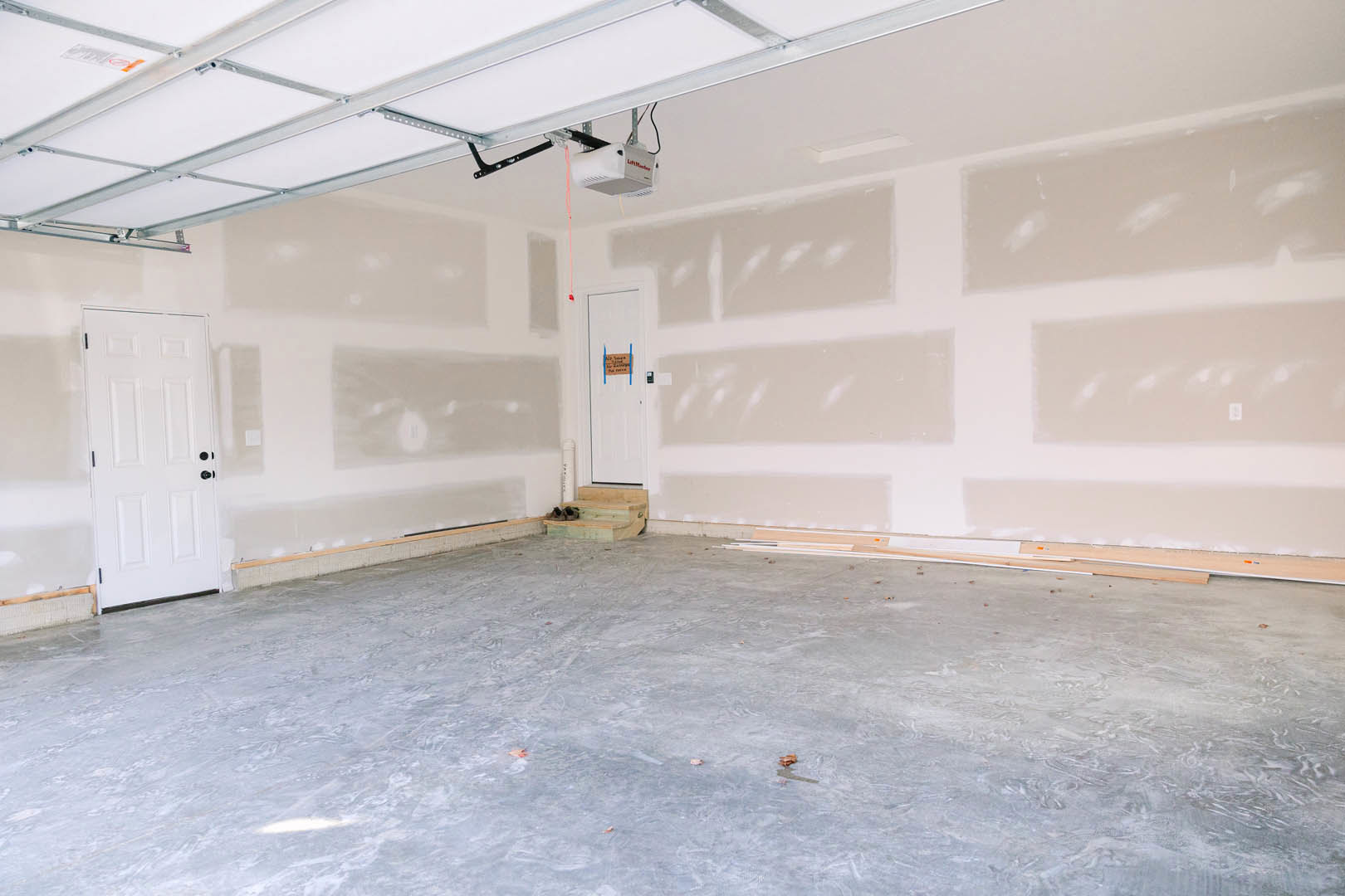 White plaster wall and ceiling, white door with black knobs, concrete floor scattered with wood pieces, small sign on door, white ceiling-mounted box.