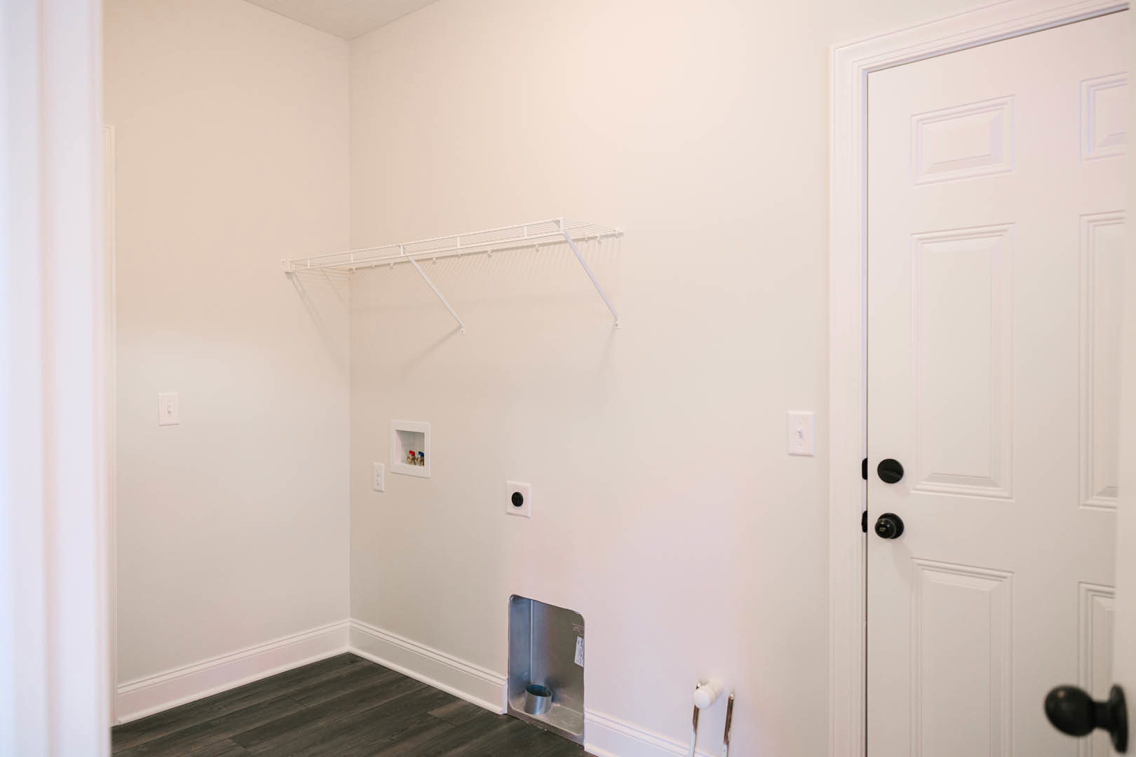 White plaster wall with floating white shelf, matte black door, wood floor with white trim, silver container on countertop, exposed metal pipe in corner, white object resting on