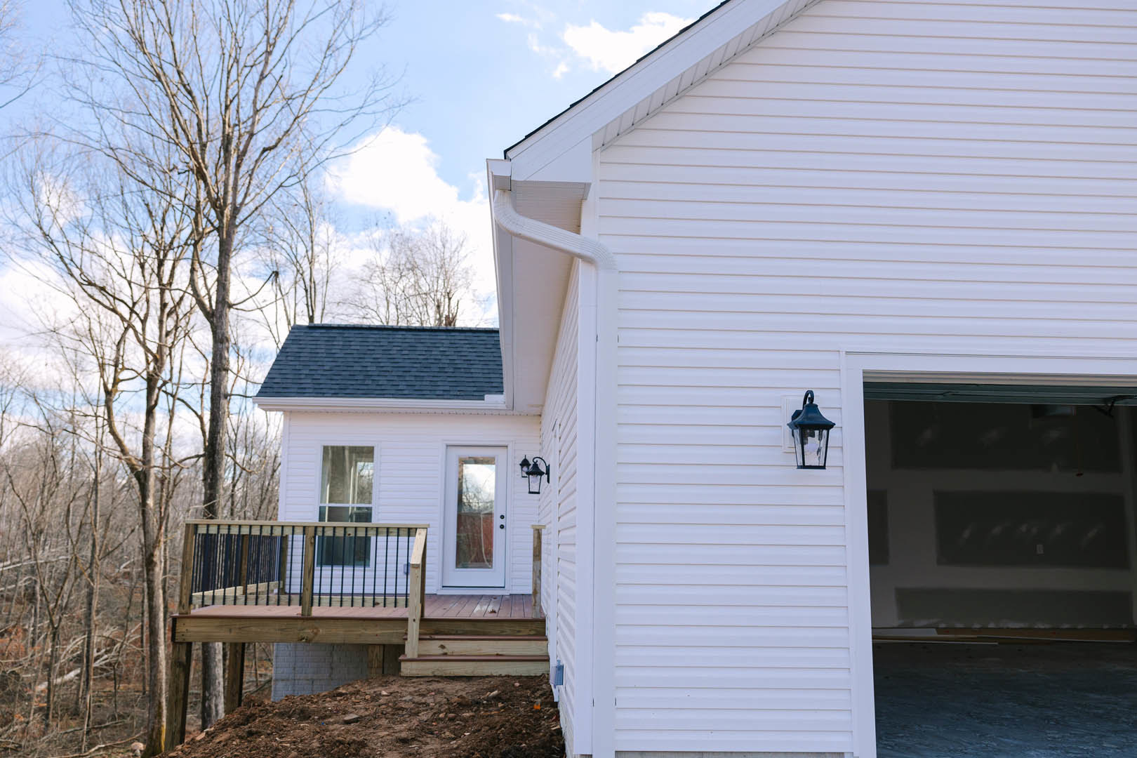 White siding house with attached garage, covered front porch, elevated wooden deck, metal railing, large windows, surrounded by trees under blue sky