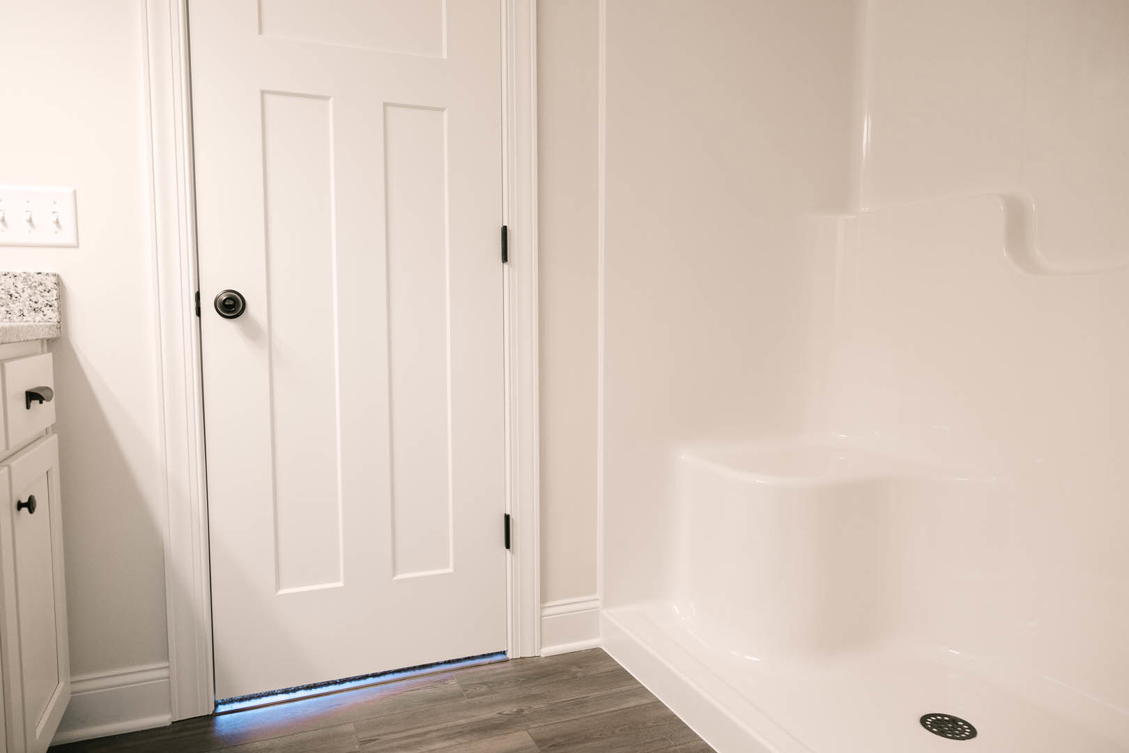 White bathroom featuring a corner shower stall with tile walls, white door with black knob, row of switches on the wall, and a close-up of a countertop.