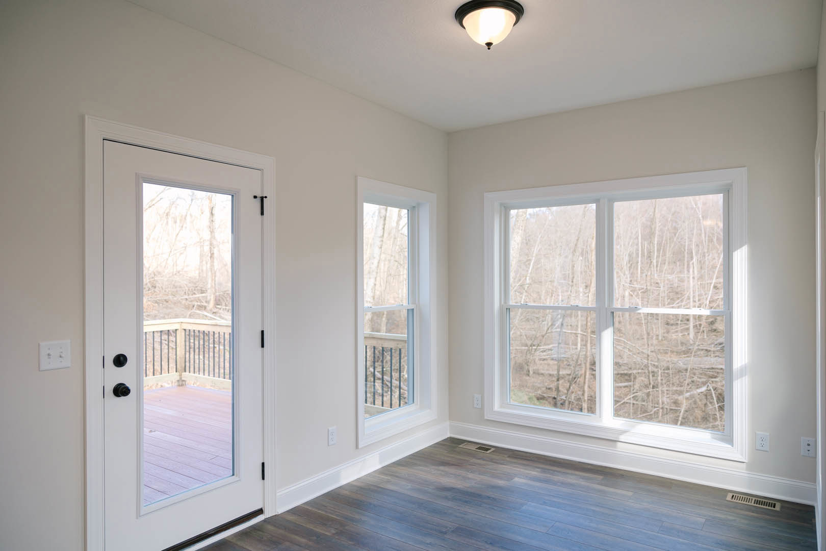 Open room with wood flooring, large windows overlooking trees, glass door leading to deck with railing, white wall with double light switch, ceiling light fixture, floor vent.