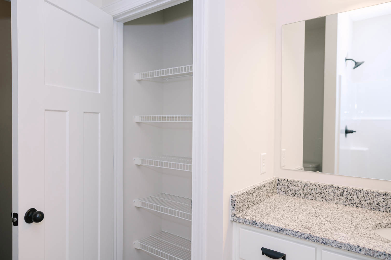 Walk-in closet with built-in white shelves, large wall mirror, and black and white speckled countertop adjacent to bathroom with white tile and chrome shower head
