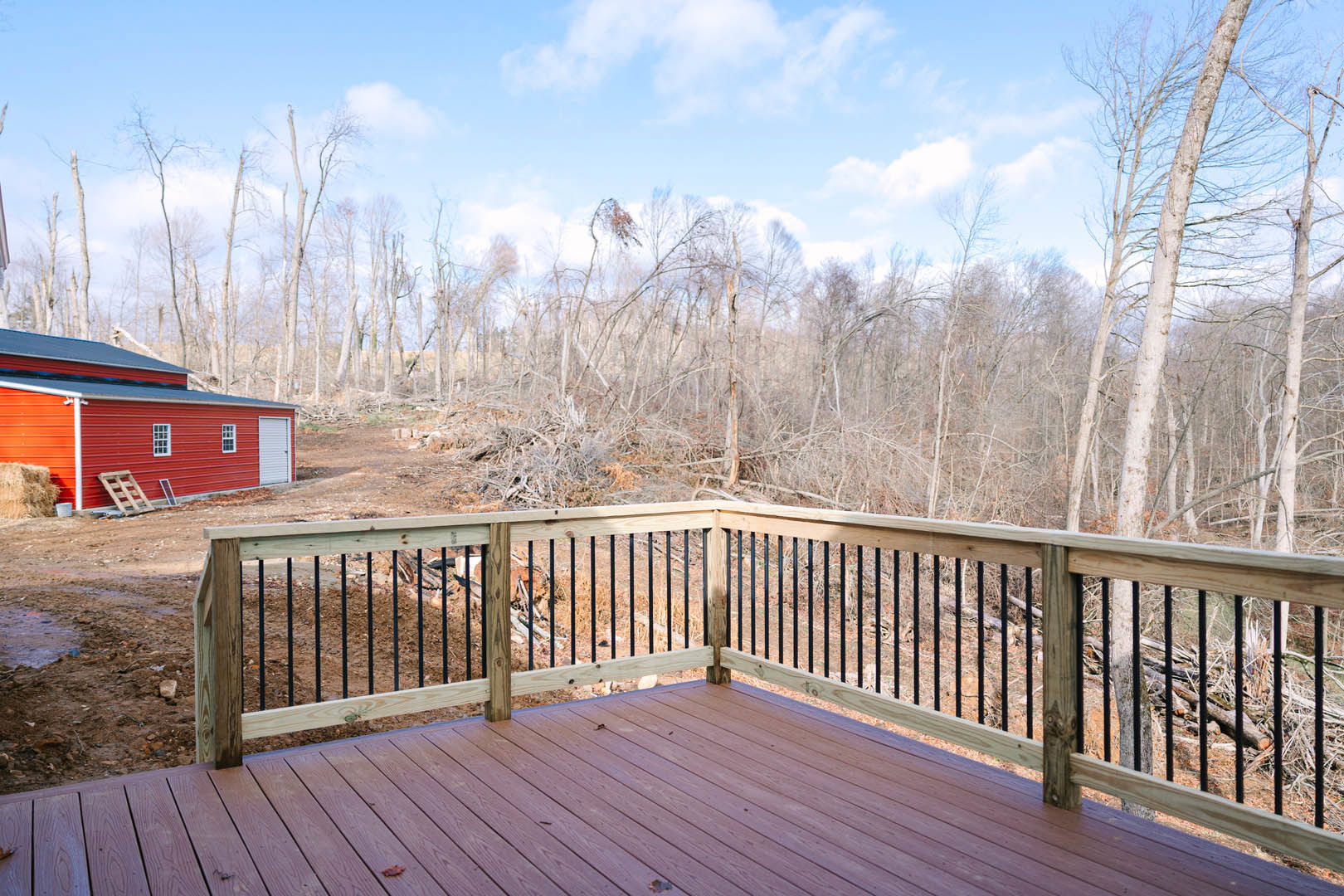 Wooden deck with black railing, red shed and ladder in background, blue sky with clouds, trees, and stack of hay nearby