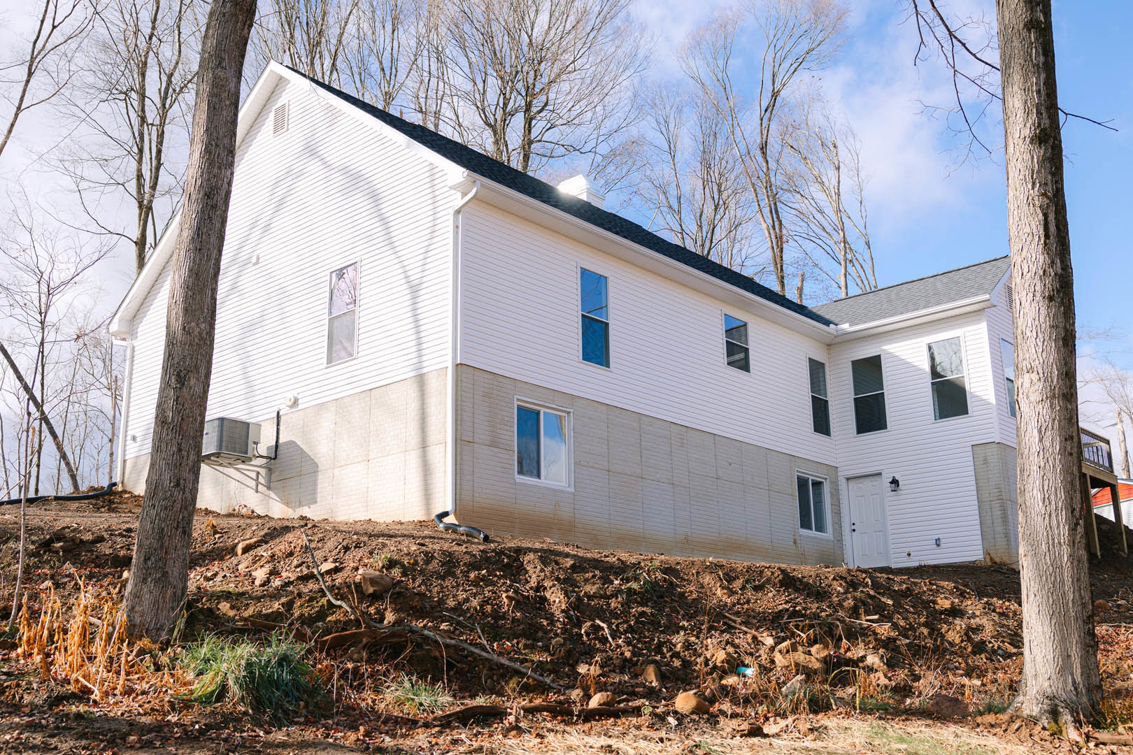Two-story home with white siding and large windows, set against a dirt hill with scattered trees and a blue sky; black pipe visible on hillside near construction area.