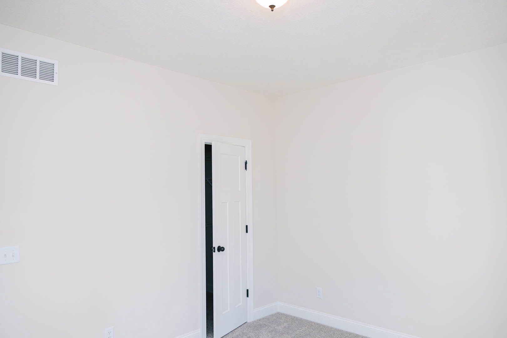 White walls and ceiling with recessed light fixture, white door with brushed metal handle, light switch and air vent on adjacent wall, smooth plaster finishes, light-colored