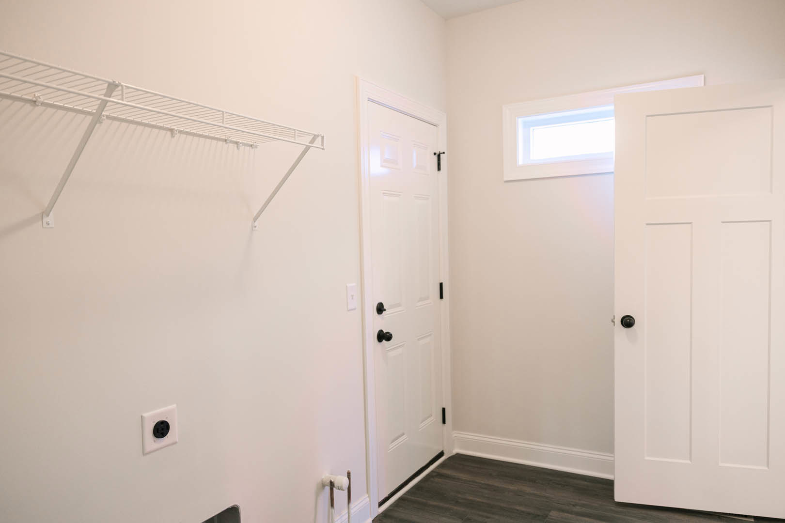 White paneled door with black knob, white floating shelf mounted on light gray wall, electrical outlet below shelf, partial view of window frame