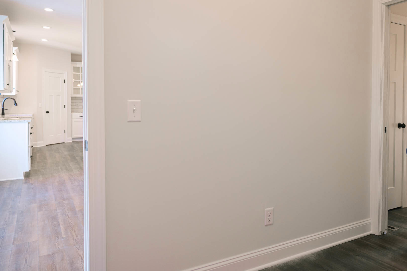 White plaster wall with a light switch, white door featuring black knobs, and partial view of flooring in a modern interior room