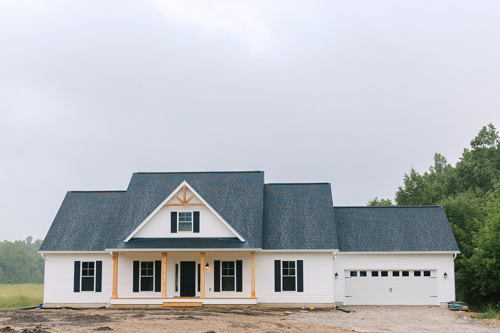 White siding house with black shingle roof, attached garage, white-framed windows, black front door with white trim, surrounded by trees under partly cloudy sky