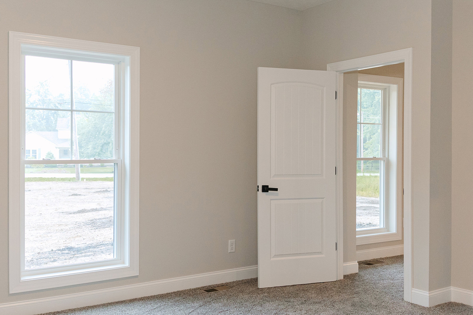 White paneled door with black handle set in a light-colored room, adjacent to a window showing a house and grassy field outside; vent and wood flooring visible.