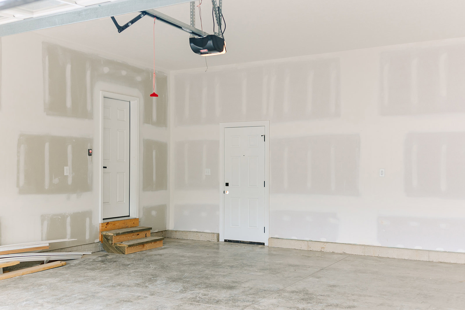White paneled door with black handle set in plaster wall, adjacent to metal shelf with bracket, wooden staircase, and window; light fixture mounted beside door, composite flooring