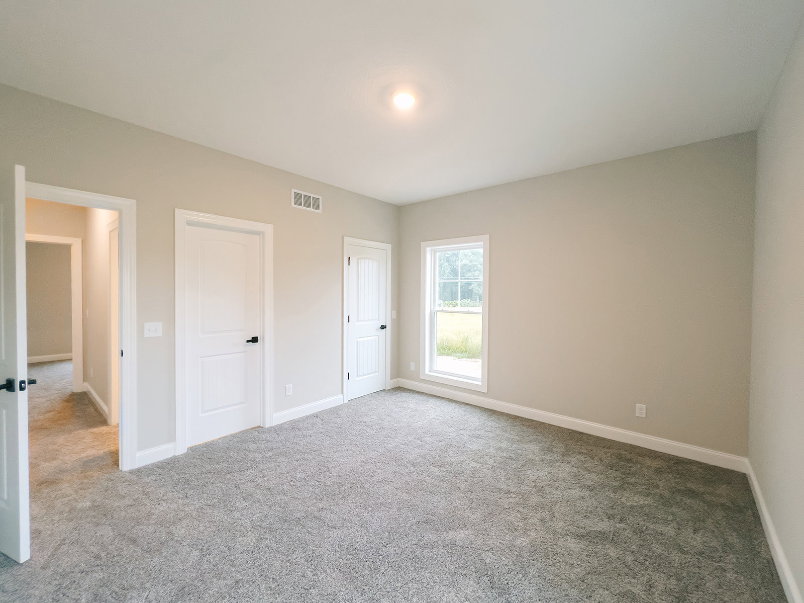 White-walled room with light carpet flooring, large window showing outdoor view, and white door featuring black handle