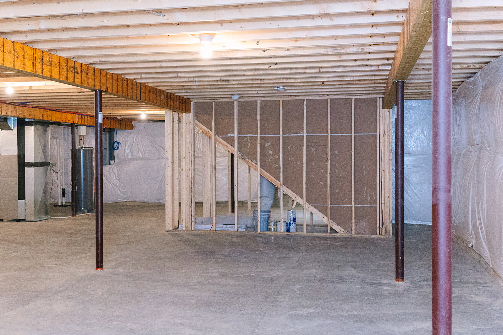 Open basement room with exposed steel beams, composite ceiling panels, white walls, staircase, and a bed covered with a white sheet; single light bulb fixture overhead, blue light