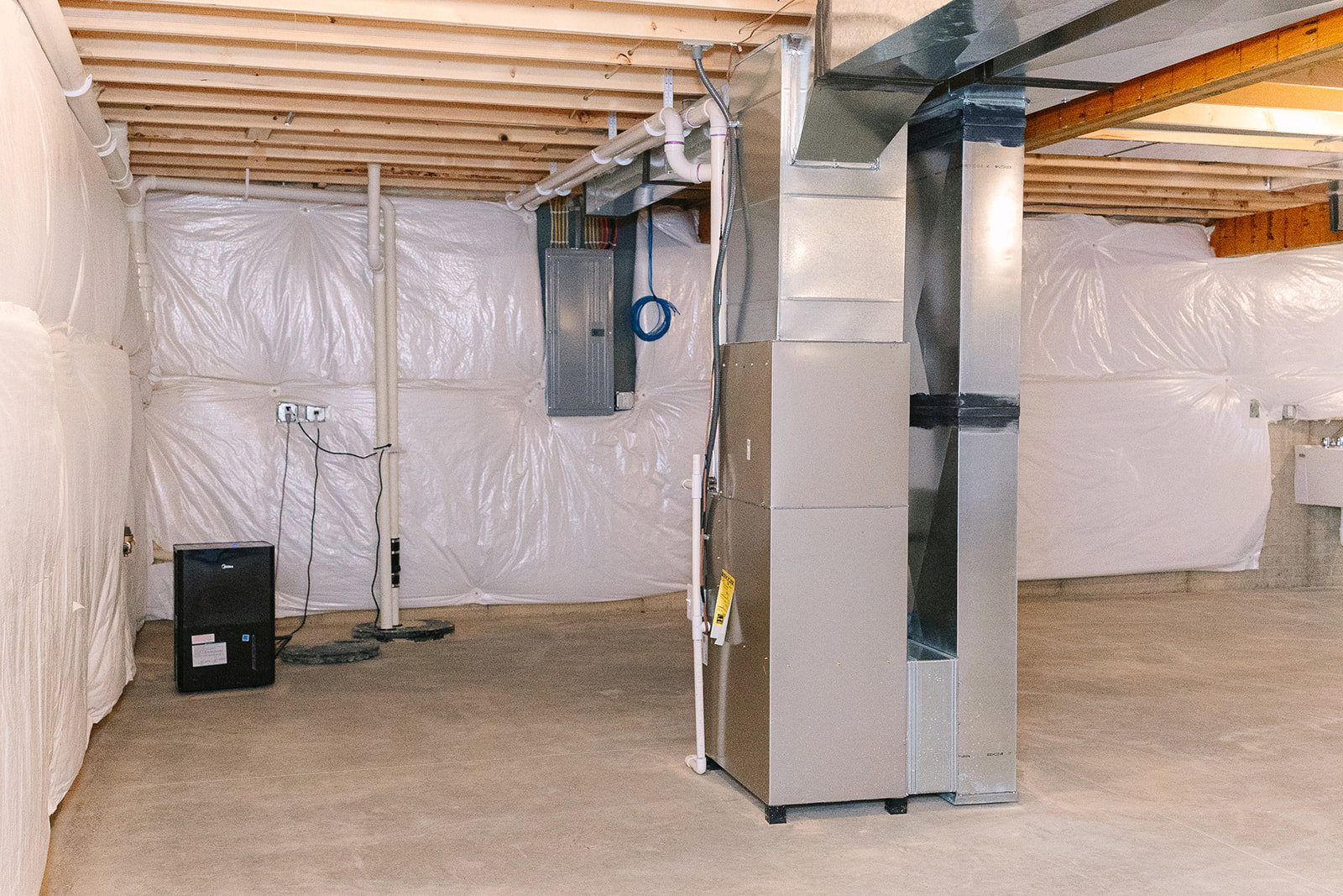 Basement room with exposed wooden ceiling beams, large black metal HVAC unit with visible pipe, blue wire on white wall, and white plastic wall covering.