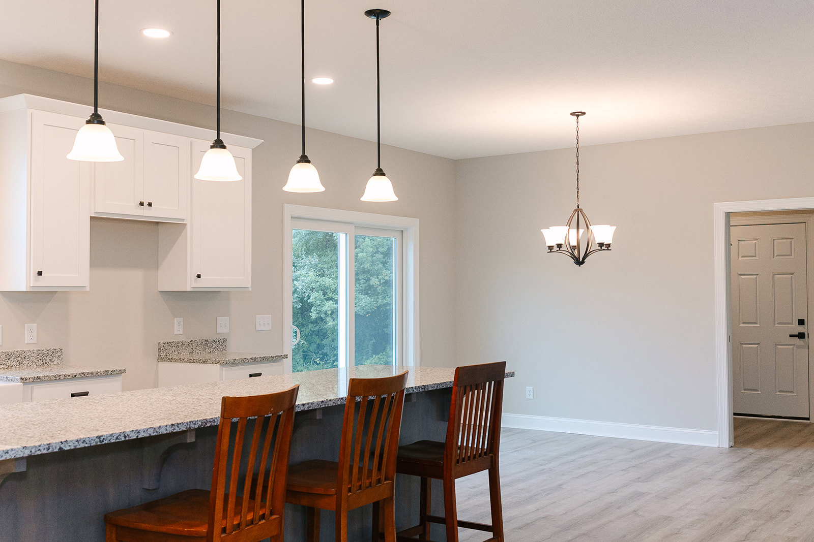 Open kitchen with light wood cabinetry, white stone countertops, bar seating with two wooden chairs, black hardware on white door, modern chandelier overhead, hardwood flooring