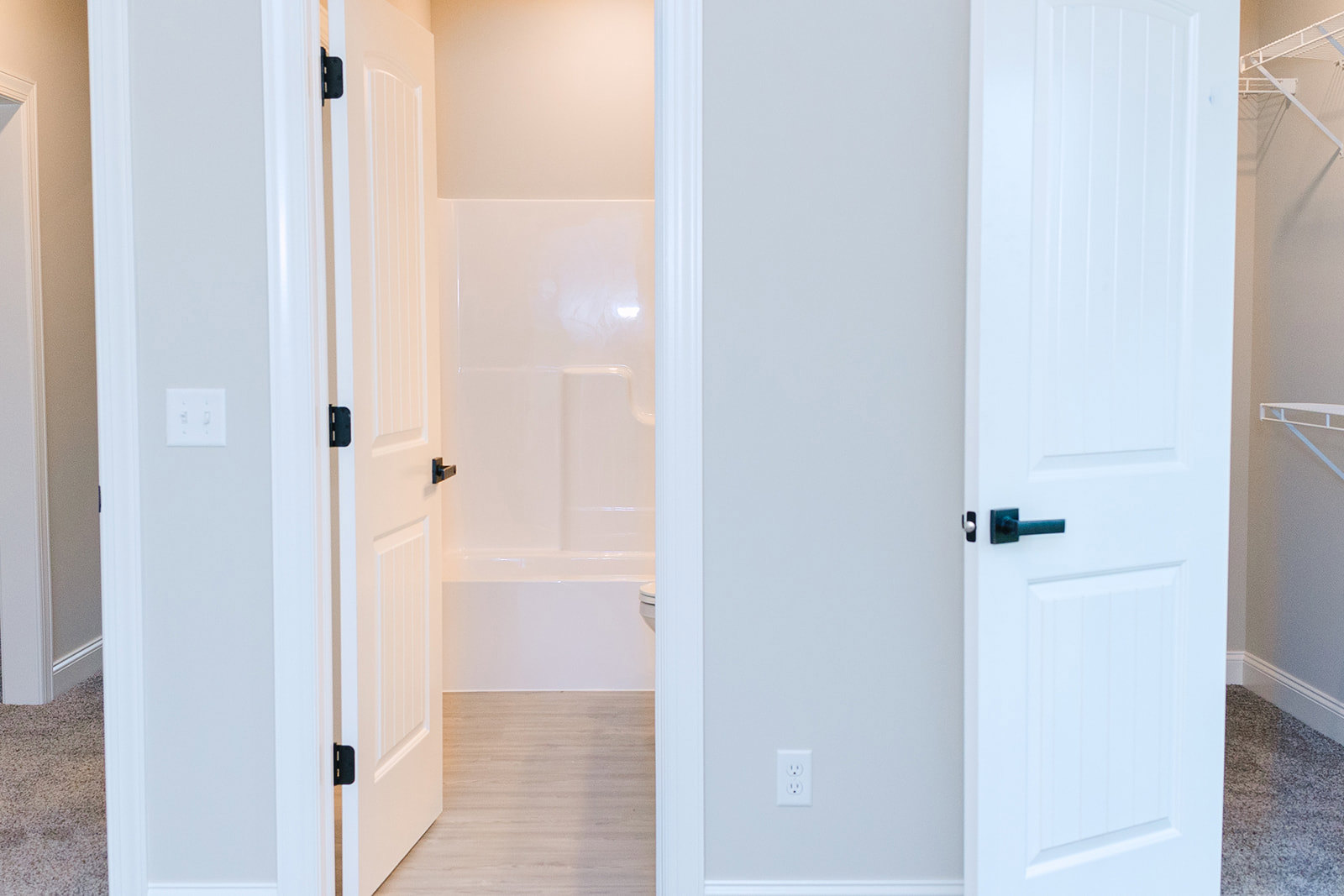 Bathroom with white walls, white wooden floor, white door featuring black handle, white outlet, and white light switch with black buttons