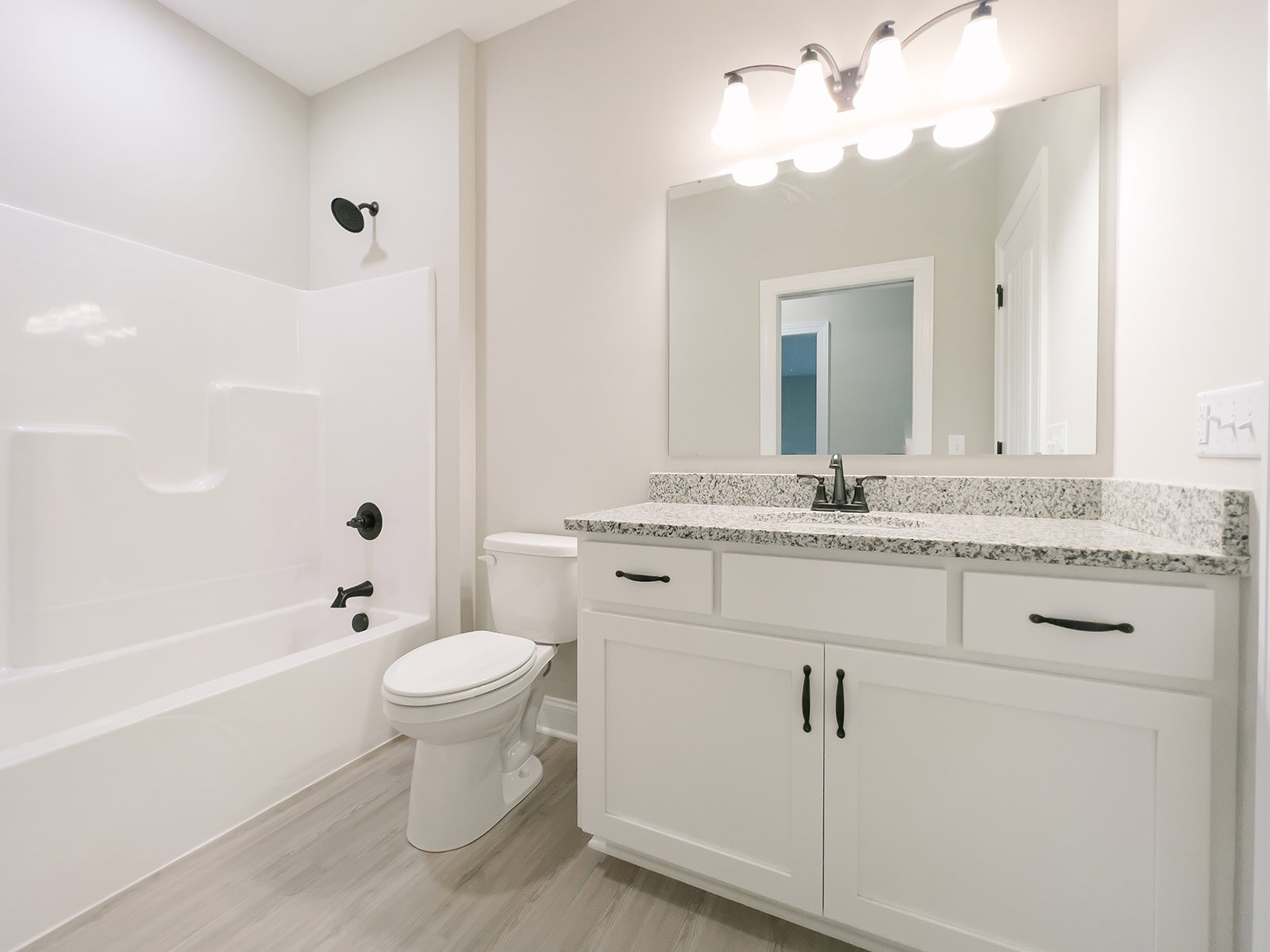 Bathroom with white toilet and bathtub, black curved shower head, white door and frame, light fixture illuminated, neutral tile walls and flooring