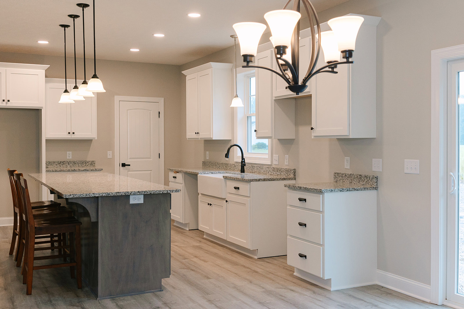 White kitchen with marble countertops, kitchen island with stools, white cabinets and drawers, close-up chandelier, white door with black hinges, hardwood flooring