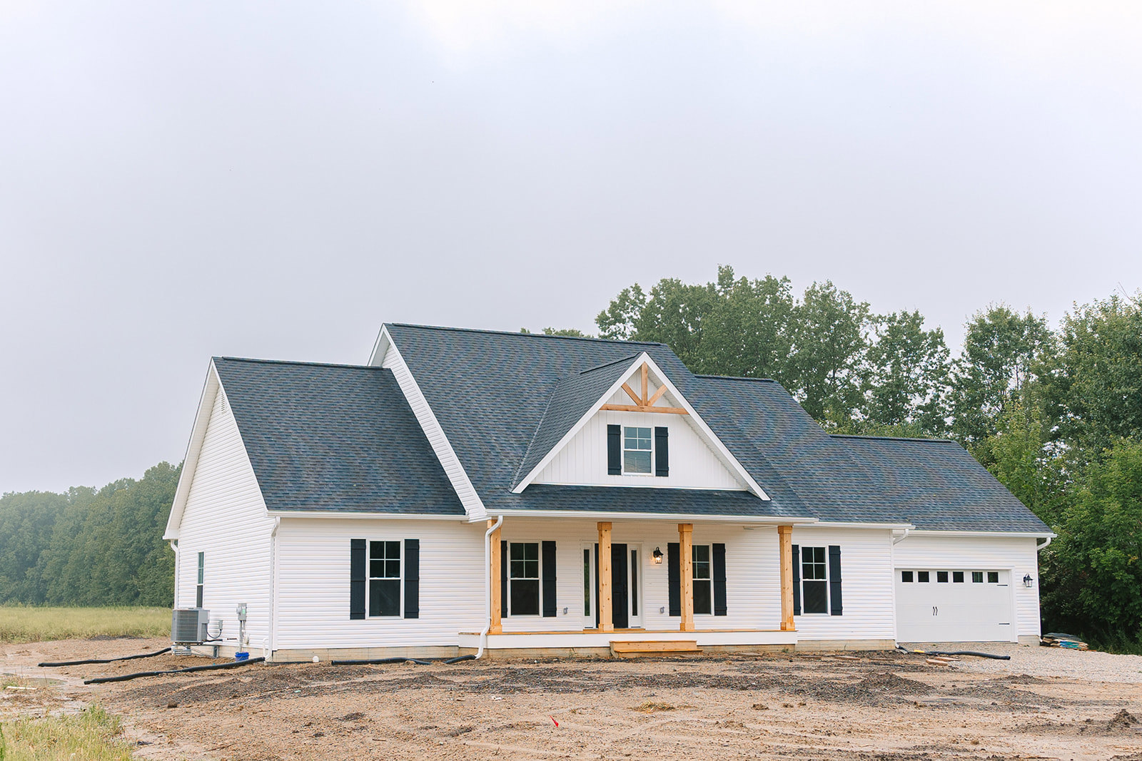 White farmhouse with black roof, attached garage, white-framed windows, and red construction equipment on dirt driveway