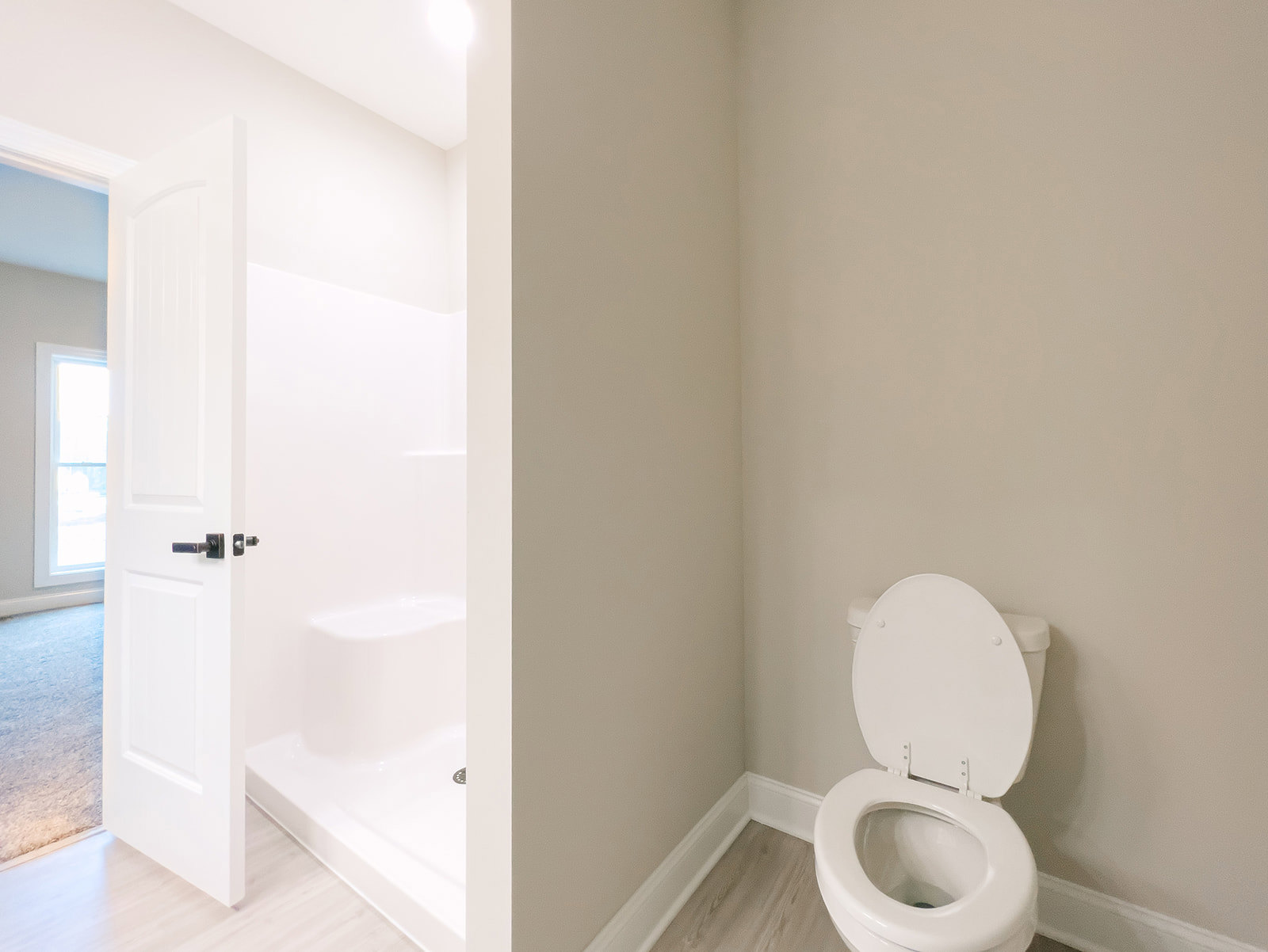 Modern bathroom with white toilet seat open, glass shower enclosure, light gray tile walls, and window letting in natural light