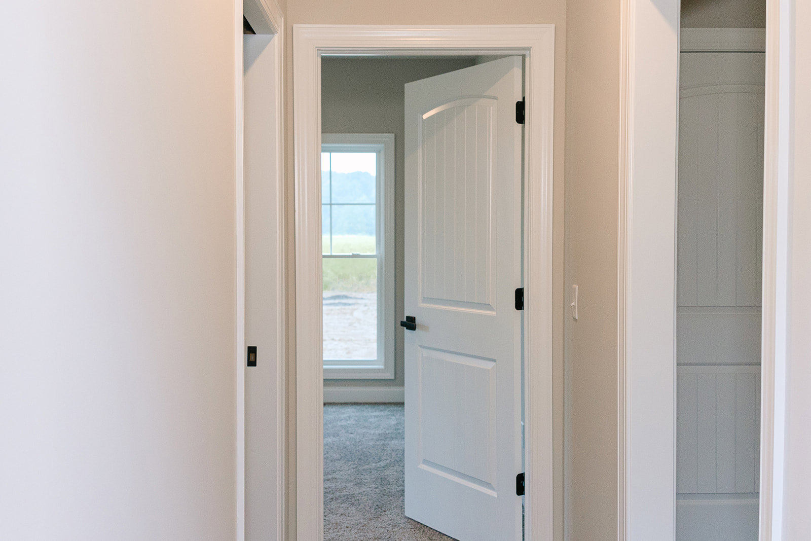 White paneled wall with open white door featuring black handles, window overlooking grassy field, light wood flooring