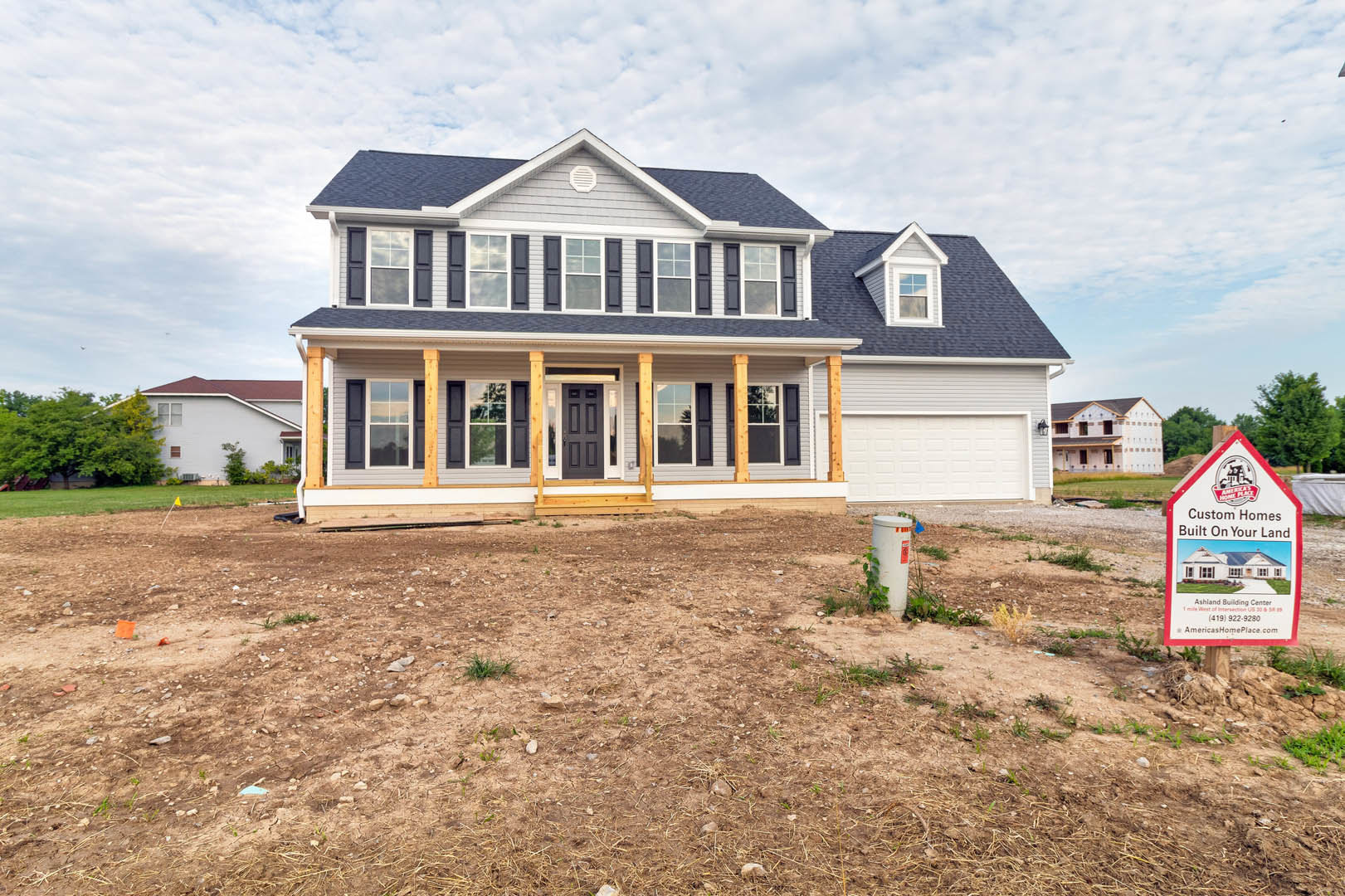 Two-story house with wide front porch, large wooden front door, expansive grassy yard, mature trees, and real estate sign near white post.