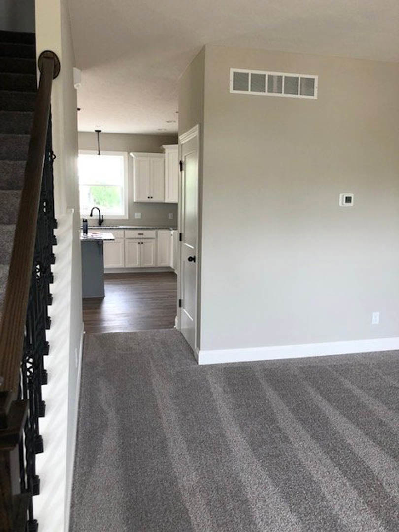 Hallway with grey carpet and white trim, white door with black handle, staircase featuring black railing, window with white frame, wall vent centered on plaster wall