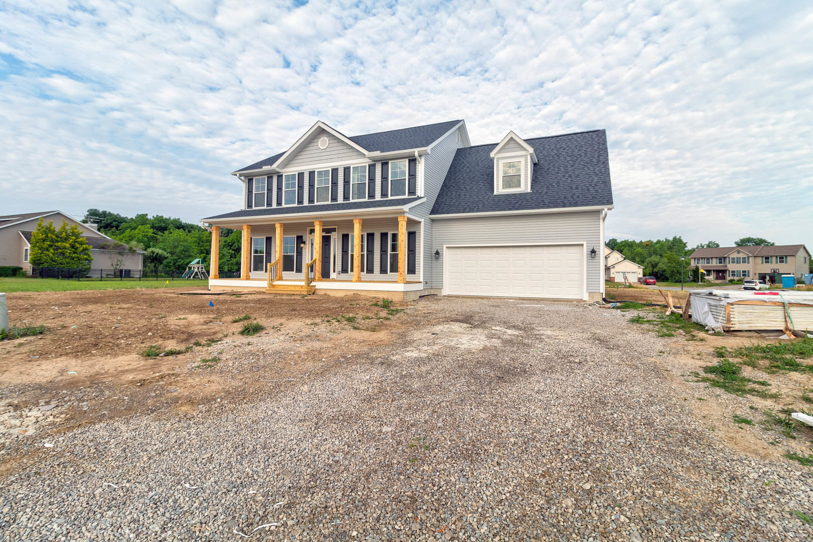 Two-car garage with white doors, gravel driveway, mature tree in front yard, several cars parked, blue sky with scattered clouds, windows along beige exterior
