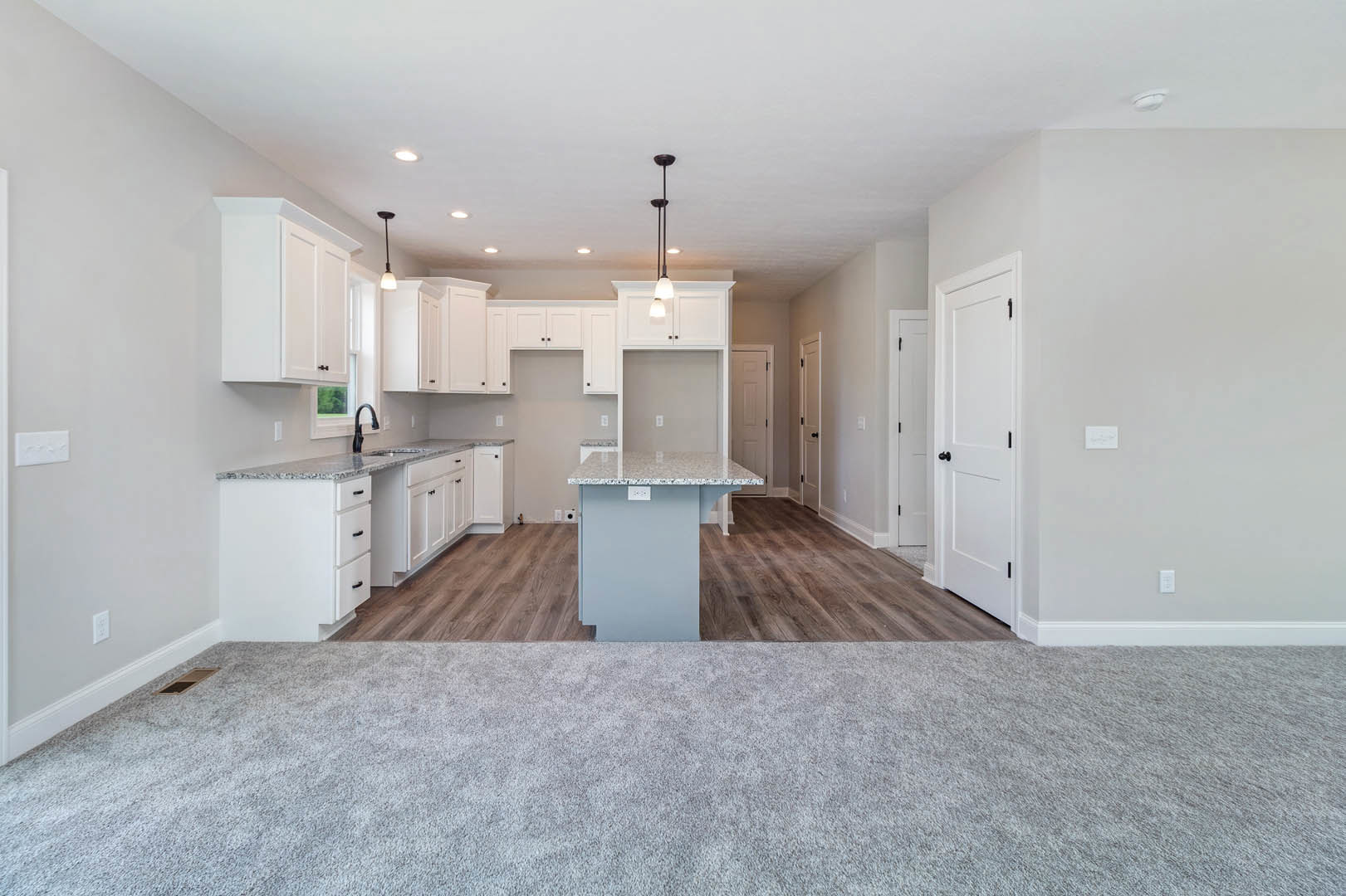 Open-concept kitchen and living room featuring white cabinetry, light gray countertops, carpeted flooring, white doors with black knobs, and a white rectangular object with text on