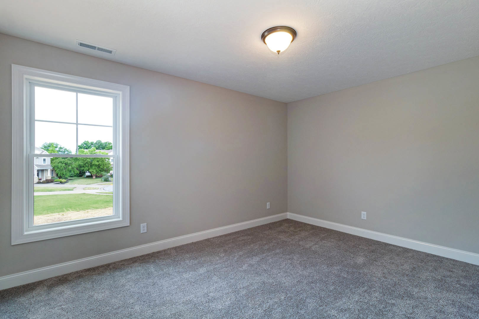 Neutral-toned carpeted room with white walls, large window overlooking neighboring house and trees, ceiling-mounted light fixture