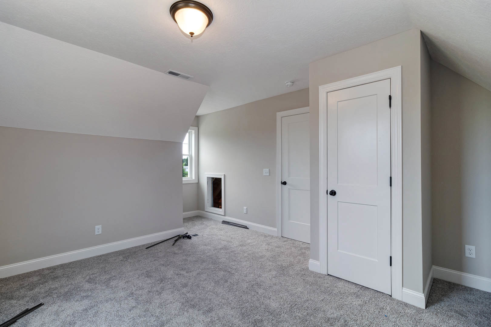 White-walled room with light carpet flooring, white doors featuring black knobs, ceiling-mounted light fixture, and a window with white trim.