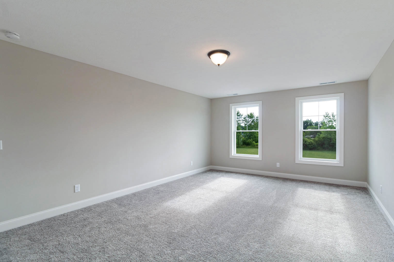 Carpeted room with two large windows, white walls, ceiling light fixture, and trees visible outside
