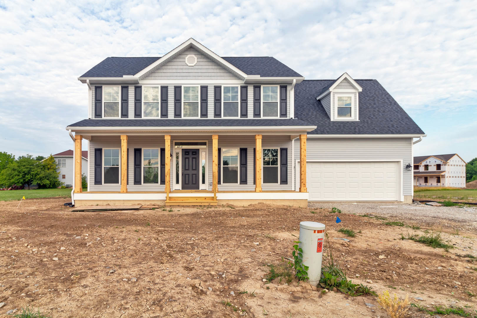 White house with yellow trim, spacious front porch with white columns, attached garage, dirt yard with small plant, mature trees in background, partly cloudy sky