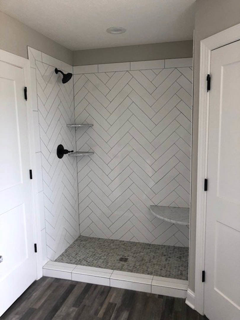 Modern bathroom shower with two black shower heads mounted on a grey tiled wall, white grout lines, and matching grey tile flooring