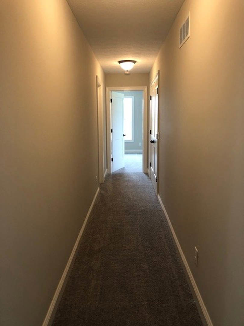 Hallway with light wood flooring, two white doors featuring black knobs and hinges, white plaster walls, ceiling light fixture, and wall sconce illuminating the space.