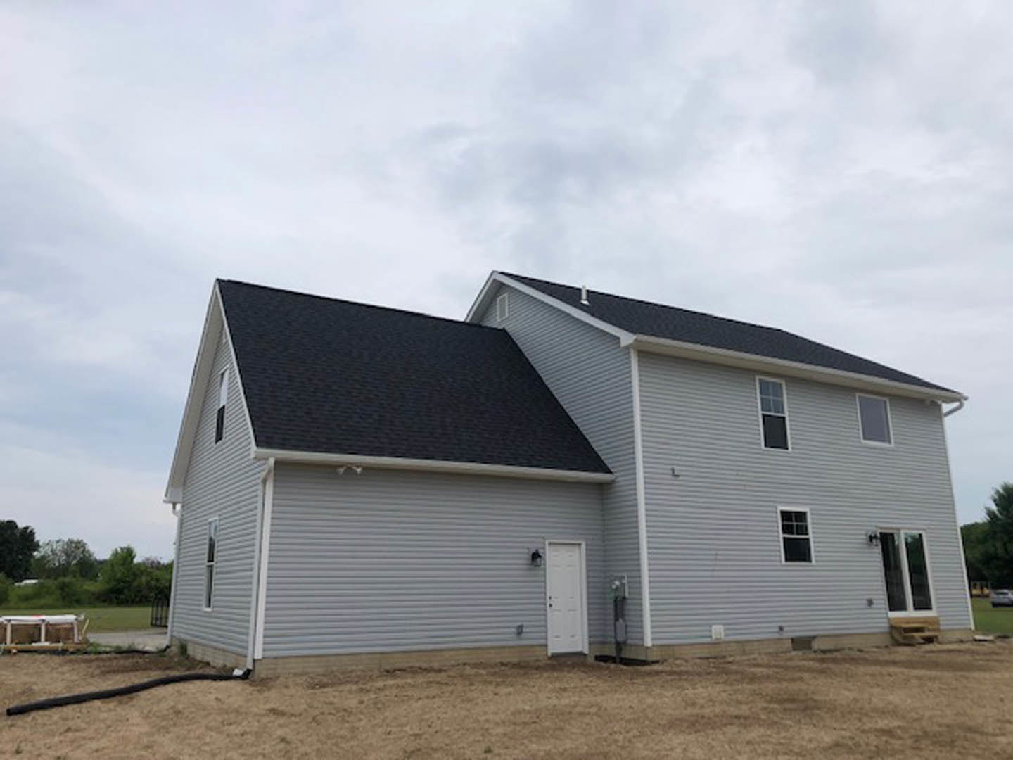 Grey siding house with black shingle roof, white front door featuring a black handle, large windows, and landscaped front yard under partly cloudy sky
