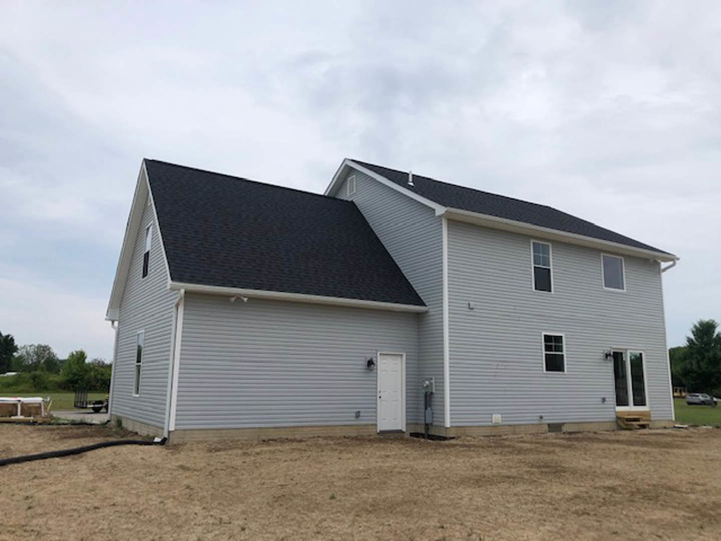Single-story house with gray siding, white-framed windows, white door with black handle, and shingled roof, set beside a dirt field under a partly cloudy sky