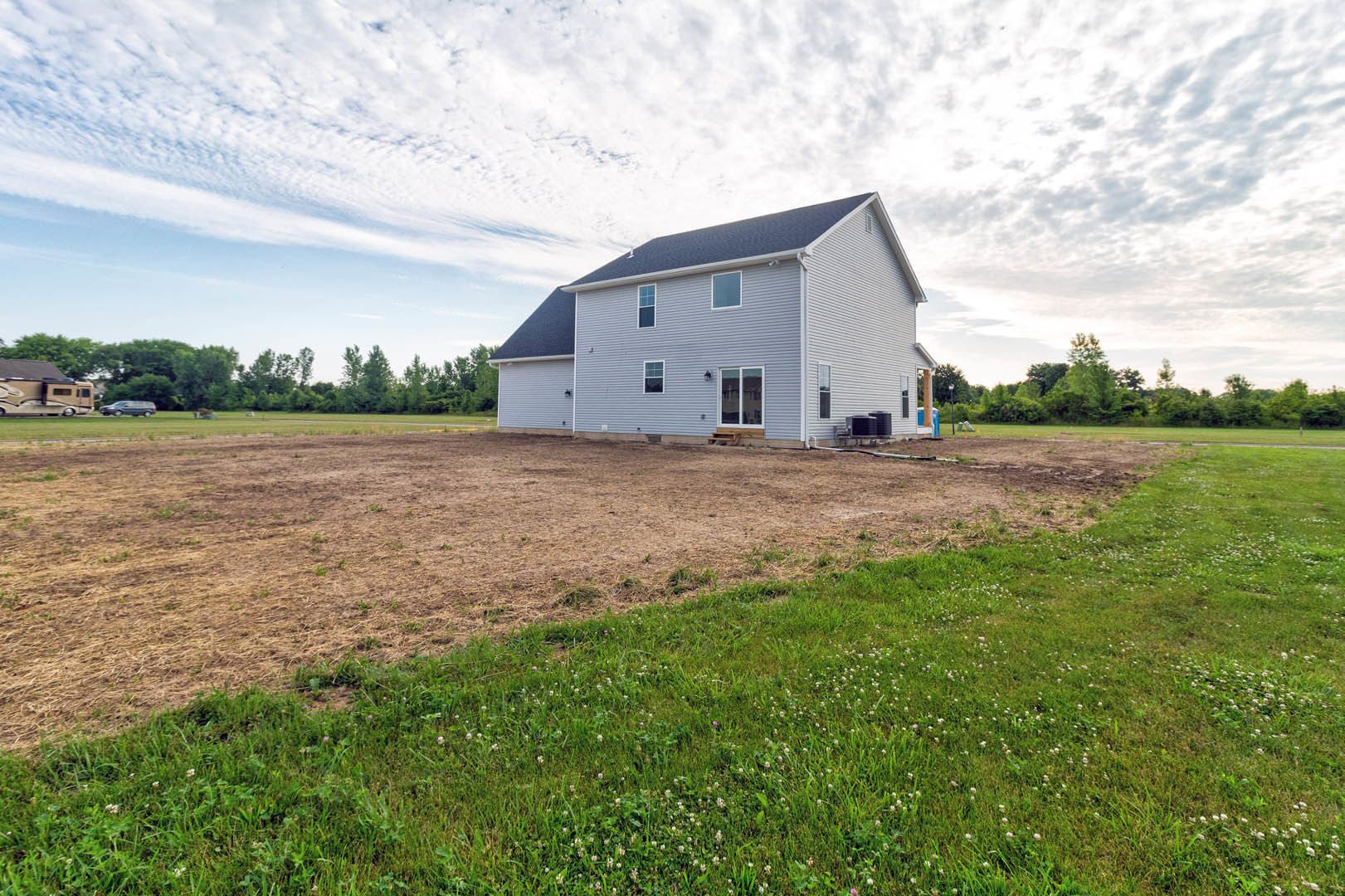 Grey house with black roof and white-framed windows, set beside a large grassy field under a cloudy sky; trees and a large RV visible near the road.