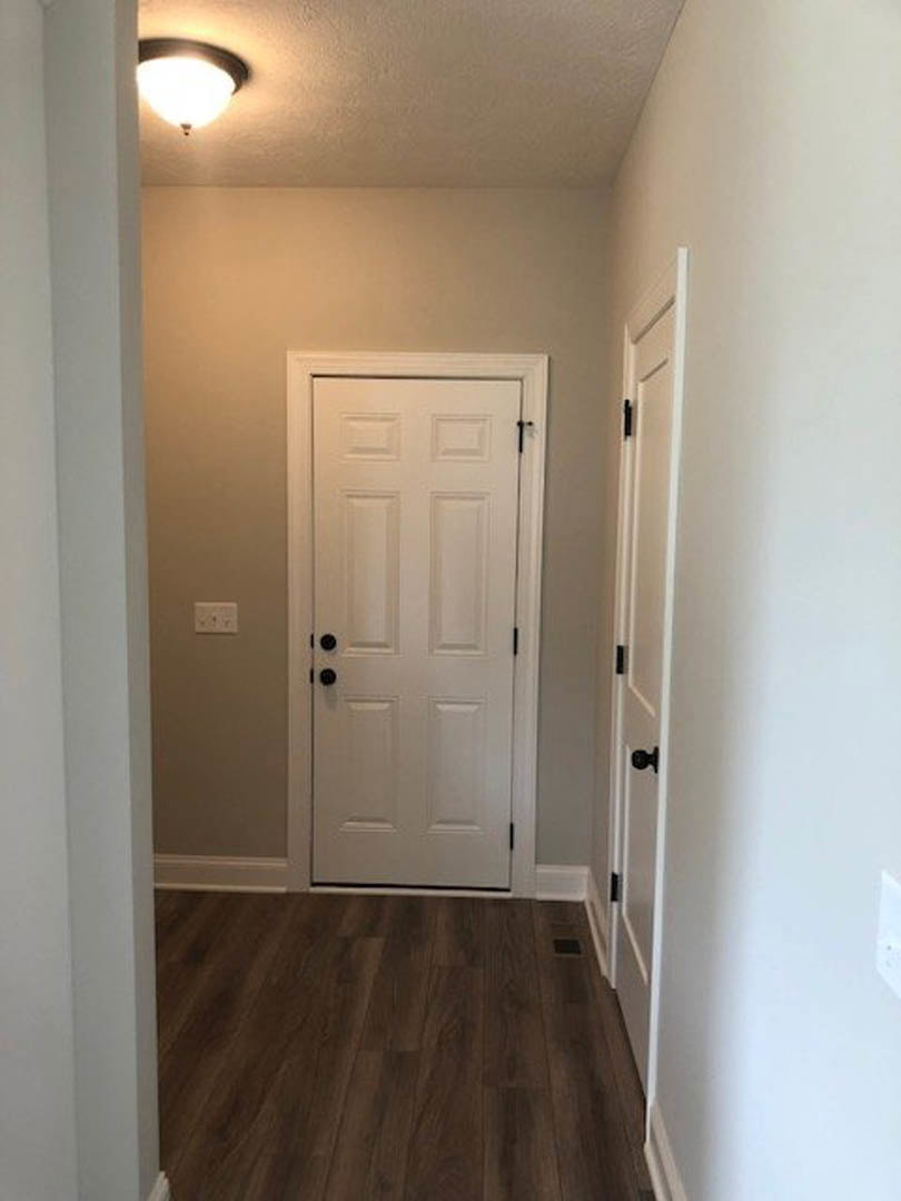 White paneled door with black handle set in a room with light wood laminate flooring, ceiling light fixture, wall vent, and white walls.