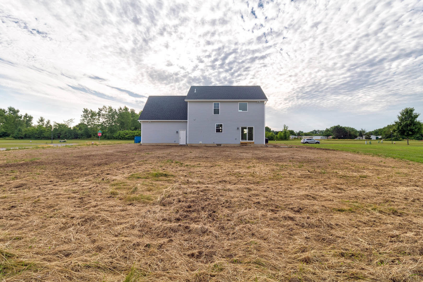 White-sided house with gabled roof, white-framed window, and white door, set beside a wide grassy field under a cloudy sky in a rural landscape.
