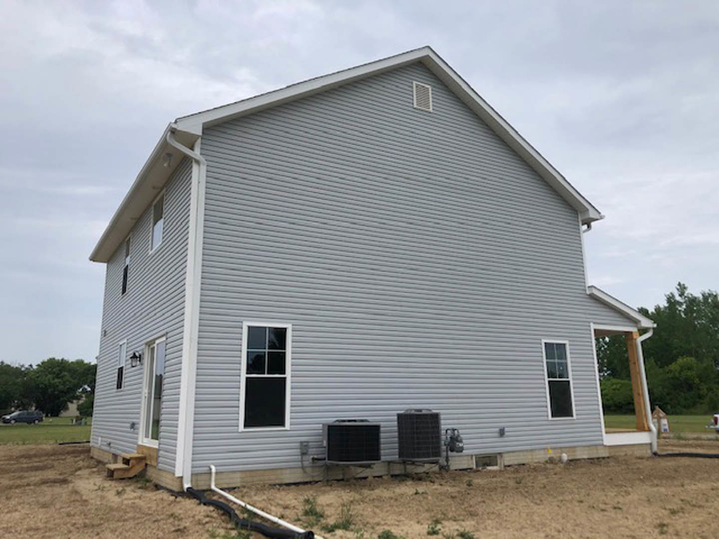 Grey siding house with white trim, multiple windows with white frames, black square object near one window, dog sitting on grassy lawn, trees and cloudy sky in background.