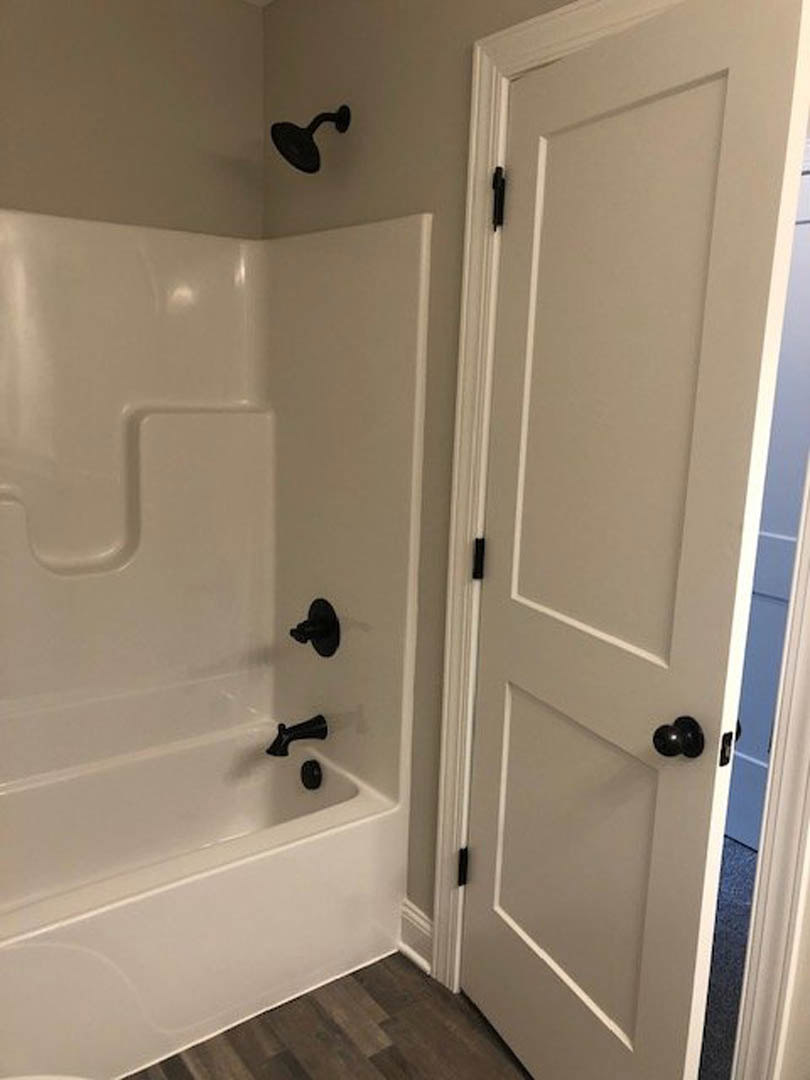White freestanding bathtub with matte black faucet, adjacent to a tiled shower wall with black shower head, wood flooring, white door with black hinges, and white walls.