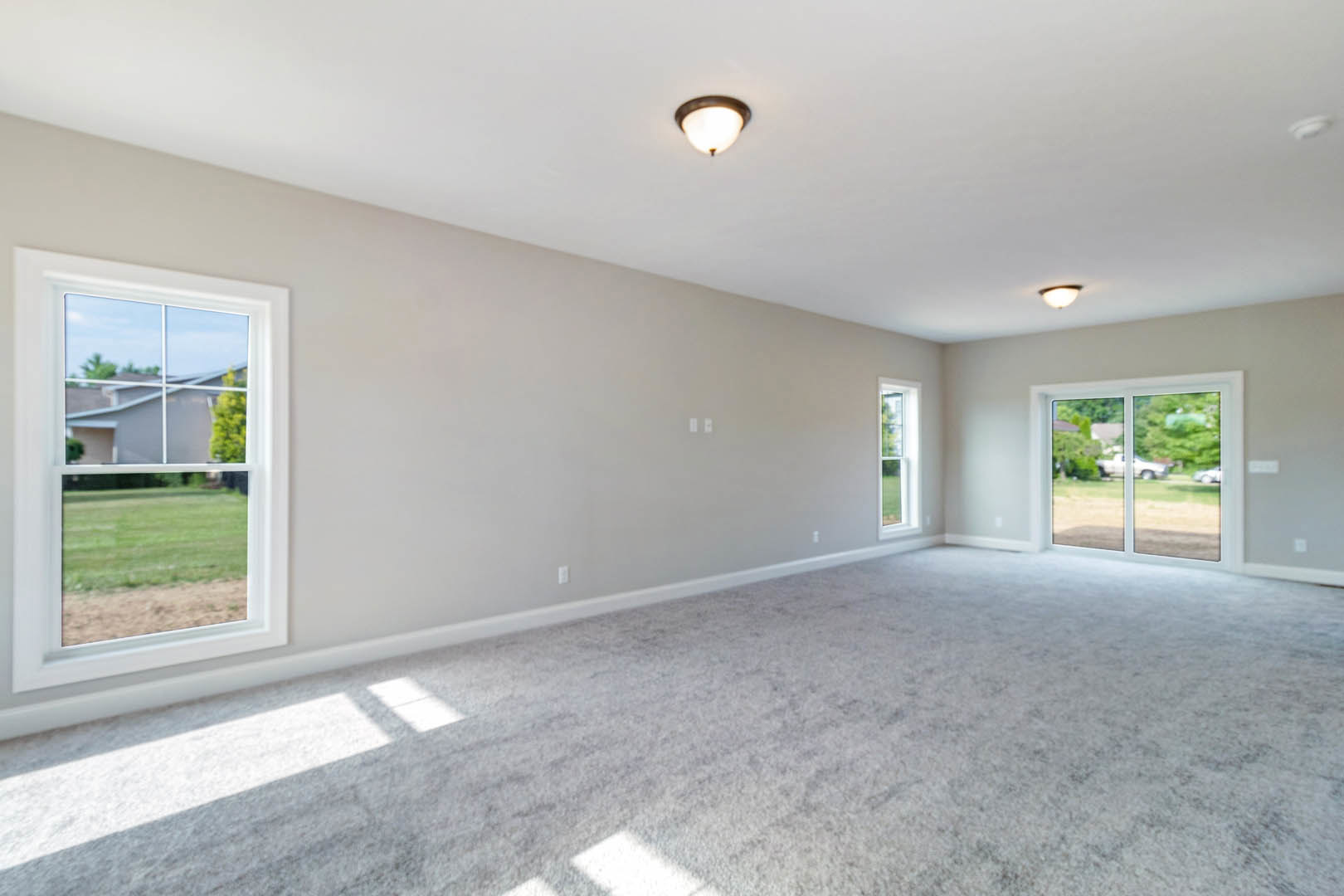 Spacious carpeted room with white walls, multiple large windows featuring white frames, ceiling light fixture, and views of a grassy yard, trees, and neighboring house